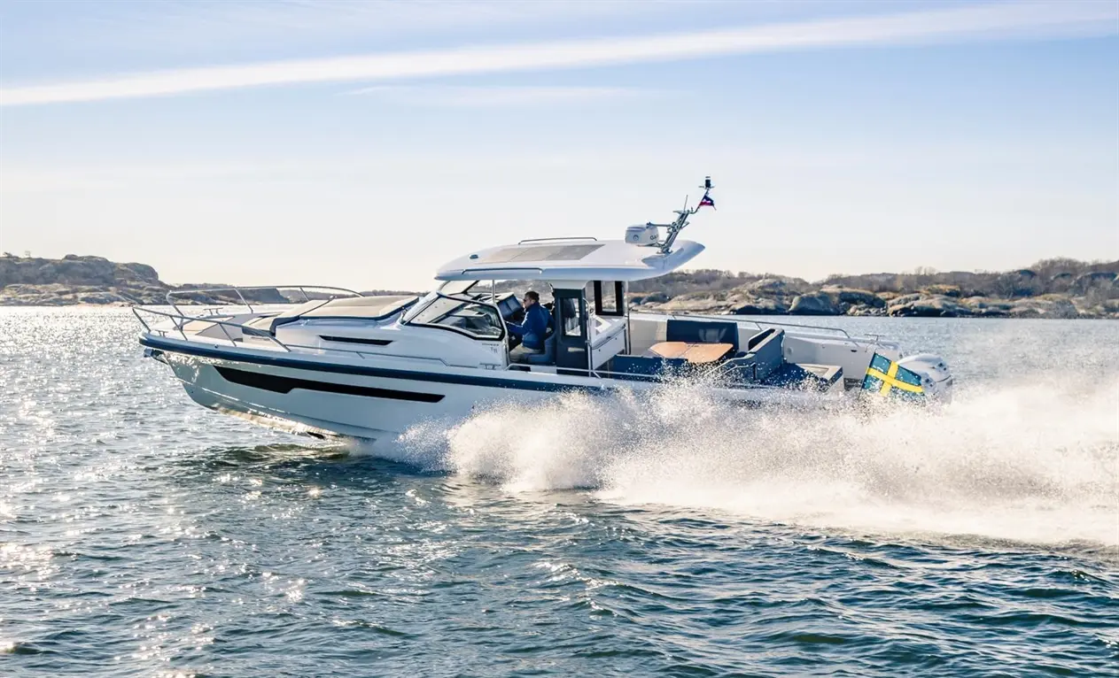 A white motorboat speeds across calm waters with a man at the helm, showcasing a Swedish flag and scenic coastline.